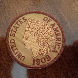 May include: A brown and gold embroidered patch with the words "United States of America" and the year "1909" in a circular design. The patch features a profile of a Native American woman wearing a headdress.