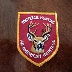 May include: Embroidered patch with a shield shape, featuring a deer head with antlers. The patch has a red background with the words "Whitetail Hunting" and "An American Heritage" in white lettering, and a yellow border.