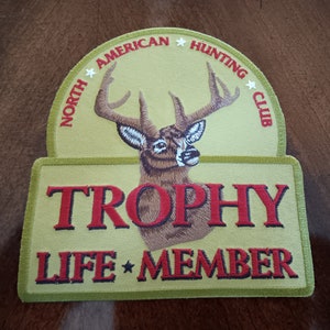 May include: A green and brown embroidered patch with a white-tailed deer and the text "North American Hunting Club" in a circle around the deer. Below the deer is the text "Trophy Life Member" in red.