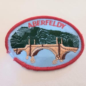May include: An oval embroidered patch featuring a light blue background with a red stitched border. The design depicts a tan stone bridge over water with dark green trees in the background. The word ABERFELDY is stitched in red at the top.