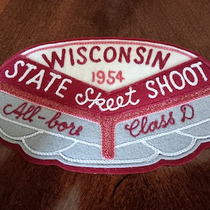 May include: A vintage felt patch with the words "WISCONSIN STATE SKEET SHOOT 1954" in white and maroon lettering. The patch has a gray and white design with the words "All-bore" and "Class D".