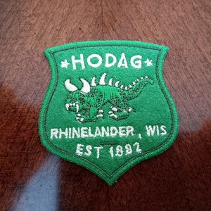 May include: A green embroidered patch with a shield shape. The patch features the word "HODAG" at the top, a green creature in the center, and the words "Rhinelander, WIS" and "EST 1892" at the bottom. The patch is on a brown surface.