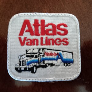 May include: White embroidered patch with red text that reads "Atlas Van Lines". A white semi-truck with blue accents is embroidered below the text.