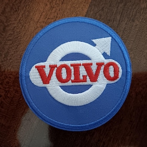 May include: A round, blue embroidered patch featuring the Volvo logo. The logo includes the word "VOLVO" in red letters, a white circle, and a white arrow pointing upwards. The patch is likely for clothing or accessories.
