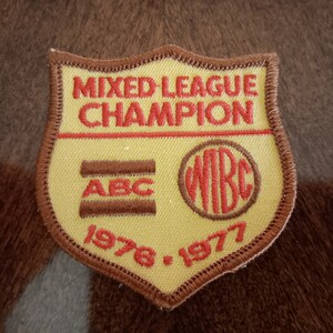May include: A yellow and brown embroidered patch with the text "MIXED LEAGUE CHAMPION" in red. The patch also features the text "ABC" and "WBC" in red, along with the years "1976" and "1977". The patch has a brown border.