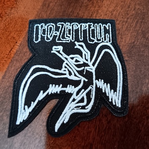 May include: Black and white embroidered patch featuring the words "LED-ZEPPELIN" above a stylized image of a winged figure. The patch has a unique shape with rounded edges and is likely designed to be sewn onto clothing or accessories.