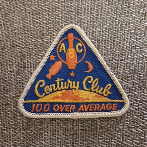 May include: A blue, triangular patch with a white border. The patch features an orange bowling pin, stars, and a crescent moon. The text "Century Club" and "100 Over Average" are in yellow and orange.