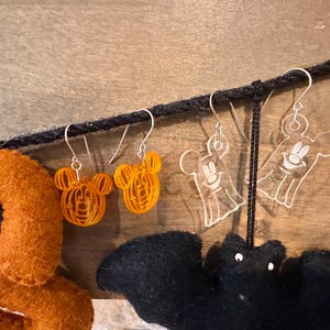 May include: Two pairs of Halloween-themed earrings. One pair features orange Mickey Mouse heads, the other pair features clear ghost figures with a smiling face.