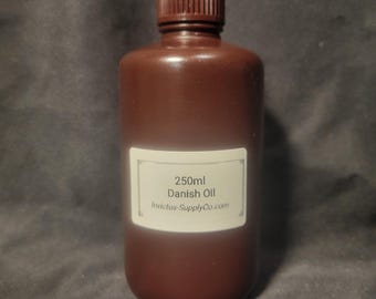 Puede incluir: Una botella marrón de aceite danés con tapa marrón. La etiqueta dice "250ml Danish Oil" y la dirección del sitio web. La botella se utiliza probablemente para proyectos de carpintería o acabado.