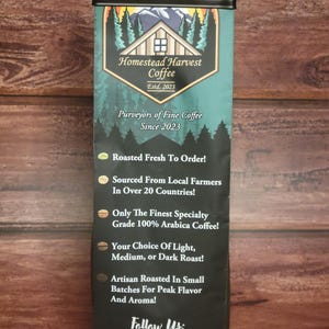 May include: A black and white bag of Homestead Harvest Coffee, established in 2023. The bag features a logo of a cabin with a mountain scene and the text "Purveyors of Fine Coffee Since 2023". The back of the bag lists the coffee's features, including "Roasted Fresh To Order!" and "Sourced From Local Farmers In Over 20 Countries!" The bag also includes a QR code with the text "Follow Us:" and "NET WT. 12 OZ".