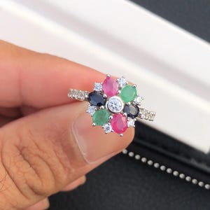 May include: A silver ring featuring a flower design with a central clear stone and surrounding petals of pink, green, and dark blue gemstones. Small clear stones are set along the band.