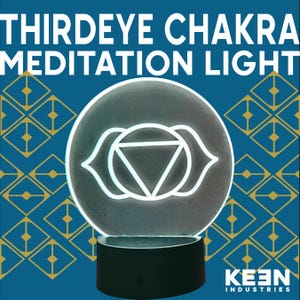 以下が含まれることがあります： 「THIRDEYE CHAKRA MEDITATION LIGHT」の文字が入ったサードアイチャクラ瞑想ライト。白いサードアイのシンボルが、黒い台座の上に置かれた円形の透明アクリルパネルに描かれています。背景は青色で、金色の幾何学模様が施されています。