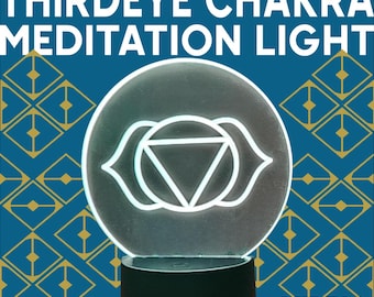 Puede incluir: Una lámpara de meditación del Tercer Ojo Chakra con el texto "THIRDEYE CHAKRA MEDITATION LIGHT". La lámpara presenta un símbolo del Tercer Ojo blanco sobre un panel circular de acrílico transparente, colocado sobre una base negra. El fondo es azul con un patrón geométrico dorado.