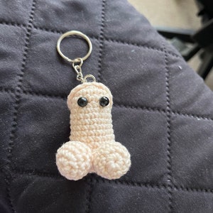 May include: A beige crocheted keychain shaped like a phallus with two black eyes. The keychain has a silver ring attached to the top. The item is made of yarn and is sitting on a black quilted surface.