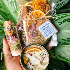 May include: A set of three items for making a simmer pot, including a glass jar filled with dried fruit and herbs, a small glass bottle with crystals and herbs, and a candle with dried fruit and crystals. The label on the jar reads "Joy & Gratitude Simmer Pot" and lists the ingredients: raw sugar, rosemary, lavender, lemon balm, nutmeg, cloves, orange, lemon, cinnamon, rose, chamomile, and calendula.