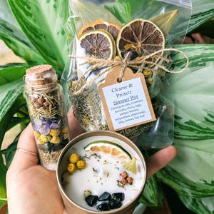 May include: A white candle with dried citrus, herbs, and black stones in a gold tin. A small glass bottle filled with herbs, crystals, and salt is next to the candle. A clear plastic bag with dried citrus, herbs, and bay leaves is behind the candle. The bag has a label that reads "Cleanse & Protect Simmer Pot" and lists the ingredients.