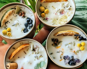 May include: Four small, round, white candles in gold tins with various dried botanicals and crystals. The candles are arranged in a circle on a hand.