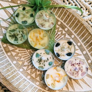 May include: A collection of eight small, round candles in metal holders, each with a unique blend of herbs and crystals. The candles are arranged on a decorative wooden tray with a carved pattern. A green ribbon and mesh bag add a touch of color.