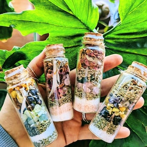 Könnte beinhalten: Vier kleine Glasflaschen gefüllt mit verschiedenen Kräutern, Kristallen und getrockneten Blumen. Die Flaschen haben Korkstopfen und sind in einer Hand angeordnet.
