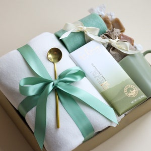 Puede incluir: Una caja de regalo con una cinta verde y una manta blanca en su interior. La caja también contiene una taza verde, una bolsa de caramelos de caramelo, una barra de chocolate negro con sal marina y una cuchara dorada.