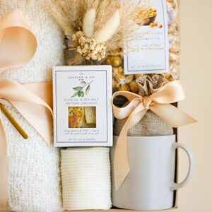 Puede incluir: Una cesta de regalo llena de una manta blanca, una taza blanca, una bolsa de palomitas de ma&iacute;z con caramelo, una caja de galletas artesanales de aceite de oliva y sal marina, y una cuchara dorada. La cesta est&aacute; decorada con una cinta beige y flores secas.