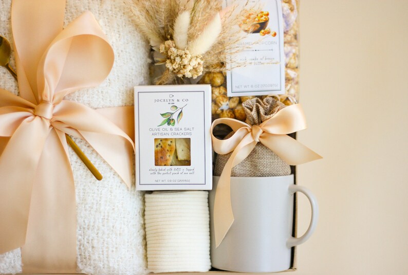 Puede incluir: Una cesta de regalo llena de una manta blanca, una taza blanca, una bolsa de palomitas de ma&iacute;z con caramelo, una caja de galletas artesanales de aceite de oliva y sal marina, y una cuchara dorada. La cesta est&aacute; decorada con una cinta beige y flores secas.