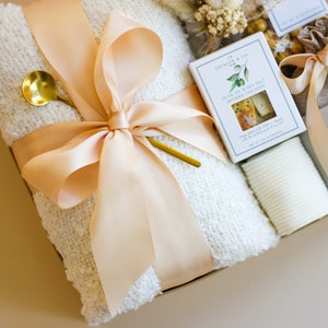 Puede incluir: Una cesta de regalo llena de una manta blanca, una taza, una peque&ntilde;a bolsa de golosinas, una peque&ntilde;a caja de galletas y una toallita blanca. La cesta est&aacute; atada con una cinta de color melocot&oacute;n.