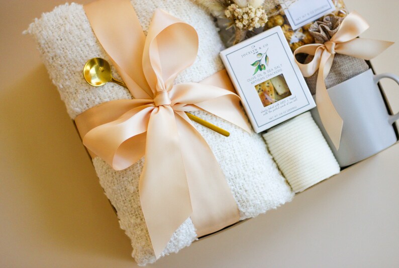 Puede incluir: Una cesta de regalo llena de una manta blanca, una taza, una peque&ntilde;a bolsa de golosinas, una peque&ntilde;a caja de galletas y una toallita blanca. La cesta est&aacute; atada con una cinta de color melocot&oacute;n.