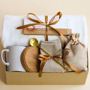 Puede incluir: Una caja de regalo de cartón marrón llena de una manta blanca, una taza blanca, un posavasos de corcho, una cuchara dorada, un par de calcetines beige, una pequeña bolsa de arpillera y una barra de chocolate con el texto "Zen Milk Chocolate" en el envoltorio.