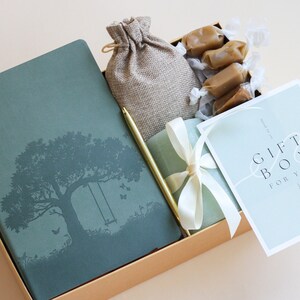 Puede incluir: Una caja de regalo llena de artículos, incluyendo un diario verde con un diseño de árbol y columpio, una pequeña bolsa de arpillera, un bolígrafo dorado, una cinta blanca atada alrededor de una caja verde y una tarjeta que dice "Gift Box for You".