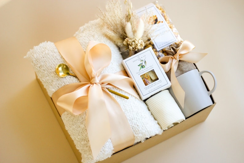 Puede incluir: Una cesta de regalo llena de una manta blanca, una taza, una bolsa de palomitas de ma&iacute;z, una peque&ntilde;a caja de galletas y un peque&ntilde;o ramo de flores secas. La cesta est&aacute; atada con una cinta de color melocot&oacute;n.