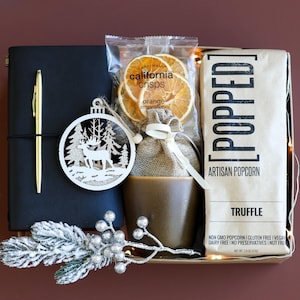 Puede incluir: Una cesta de regalo con un diario de cuero negro, un bolígrafo dorado, un adorno de madera, una bolsa de palomitas de maíz, una bolsa de chips de naranja y una taza marrón. La cesta está decorada con una rama de vegetación escarchada y una cadena de luces.