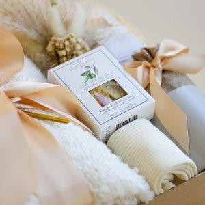 Puede incluir: Una cesta de regalo llena de art&iacute;culos acogedores, que incluyen una manta de punto blanca, un par de calcetines blancos, una taza gris y una caja de galletas artesanales de aceite de oliva y sal marina. La cesta est&aacute; atada con una cinta beige.
