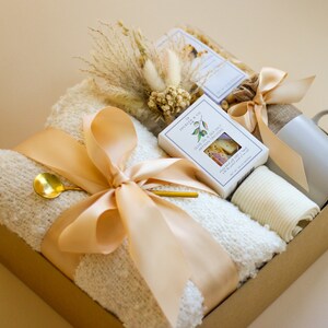 Puede incluir: Una cesta de regalo llena de una manta blanca, una taza, una peque&ntilde;a caja de galletas y una peque&ntilde;a bolsa de flores secas. La cesta est&aacute; atada con una cinta beige.