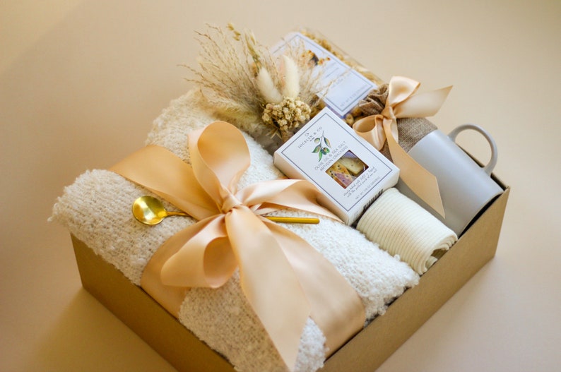Puede incluir: Una cesta de regalo llena de una manta blanca, una taza, una peque&ntilde;a caja de galletas y una peque&ntilde;a bolsa de flores secas. La cesta est&aacute; atada con una cinta beige.