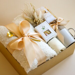 Puede incluir: Una caja de regalo marr&oacute;n llena de una manta de lana blanca, una taza blanca, una toalla de mano blanca, una peque&ntilde;a caja de jab&oacute;n blanca y una cuchara dorada. La caja est&aacute; atada con una cinta beige y decorada con flores secas.