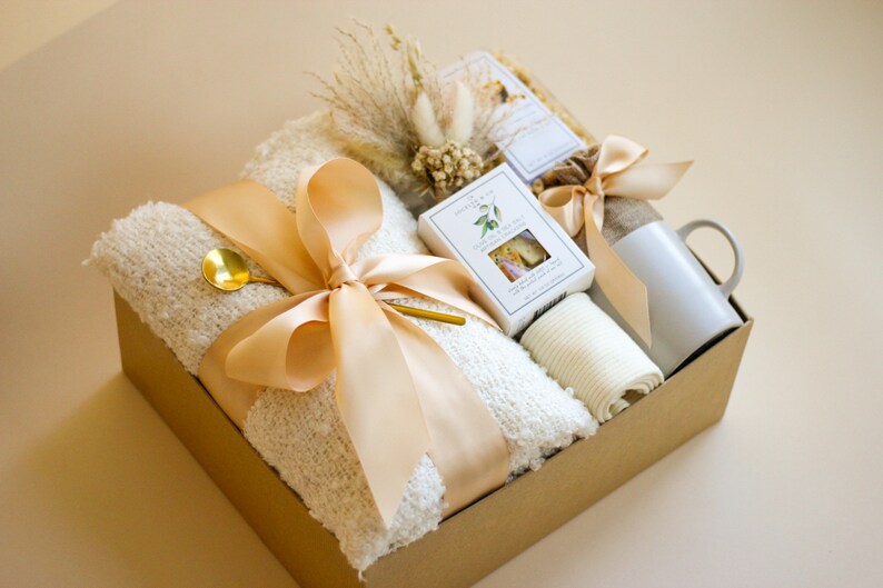 Puede incluir: Una caja de regalo marr&oacute;n llena de una manta de lana blanca, una taza blanca, una toalla de mano blanca, una peque&ntilde;a caja de jab&oacute;n blanca y una cuchara dorada. La caja est&aacute; atada con una cinta beige y decorada con flores secas.