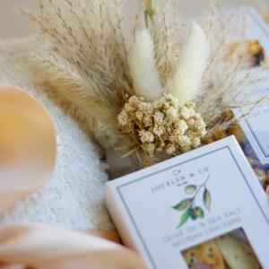 Puede incluir: Una cesta de regalo con una toalla blanca, una cinta beige, flores secas y una caja de galletas artesanales de aceite de oliva y sal marina de Jocelyn & Co.