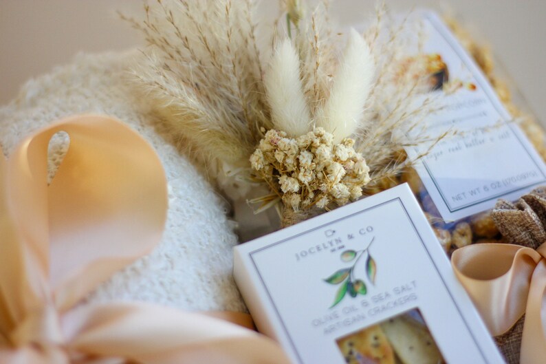 Puede incluir: Una cesta de regalo con una toalla blanca, una cinta beige, flores secas y una caja de galletas artesanales de aceite de oliva y sal marina de Jocelyn & Co.