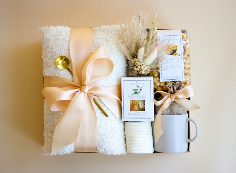 Puede incluir: Una cesta de regalo con una manta blanca, una taza, una bolsa de palomitas de ma&iacute;z, una caja de galletas, una cuchara dorada y flores secas.