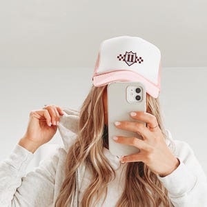 May include: A pink and white trucker hat with a burgundy shield logo with the number '11' and checkered pattern on it.