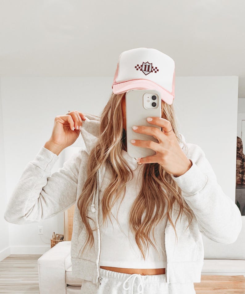 May include: A pink and white trucker hat with a burgundy shield logo with the number '11' and checkered pattern on it.