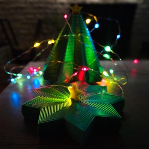 May include: A green, 3D-printed Christmas tree with a star on top, illuminated by colorful string lights. A star-shaped base with a yellow star in the center complements the tree. The festive decor is set on a dark surface.