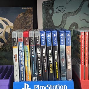 May include: A blue PlayStation game holder displaying several PS3 and PS4 game cases. Games include Call of Duty, Battlefield 1, and The Legend of Zelda. The holder has the PlayStation logo.