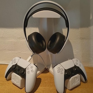 以下が含まれることがあります： 白いヘッドセットと、スタンドに置かれた白と黒のゲームコントローラー2つ。コントローラーにはPlayStationのロゴがあります。ヘッドセットは黒いイヤーカップと白いヘッドバンドを備えています。