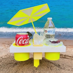 May include: A beach picnic setup with a yellow and white umbrella, a can of Coca-Cola, a bottle of water, and plastic cutlery on a white tray. The tray has yellow cup holders and is on the sand near the ocean.