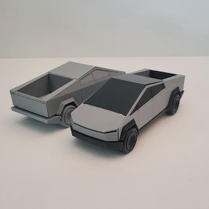 May include: Two gray toy pickup trucks with black wheels and windows are displayed on a white surface. The trucks have a geometric design and a futuristic appearance. One truck has an open bed.