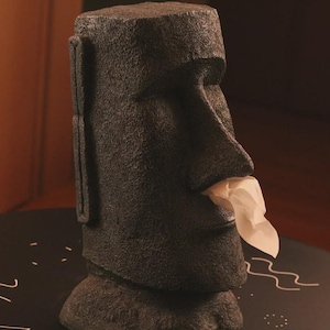 Pu&ograve; includere: Un porta fazzoletti a forma di statua Moai grigio scuro. La scultura simile a pietra ha una superficie strutturata e un fazzoletto che esce dalla bocca. Il porta fazzoletti &egrave; su una superficie nera.