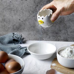 May include: A white ceramic egg separator in the shape of a chick, with a yellow beak, is held over a white bowl. Egg white is dripping from the chick's beak. Other bowls contain eggs and flour. A gray cloth is in the background.