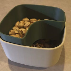 May include: A two-toned serving bowl with a green top section and a white base. The green section is designed to hold the shells of pistachios, while the white base holds the nuts. The bowl is filled with pistachios.